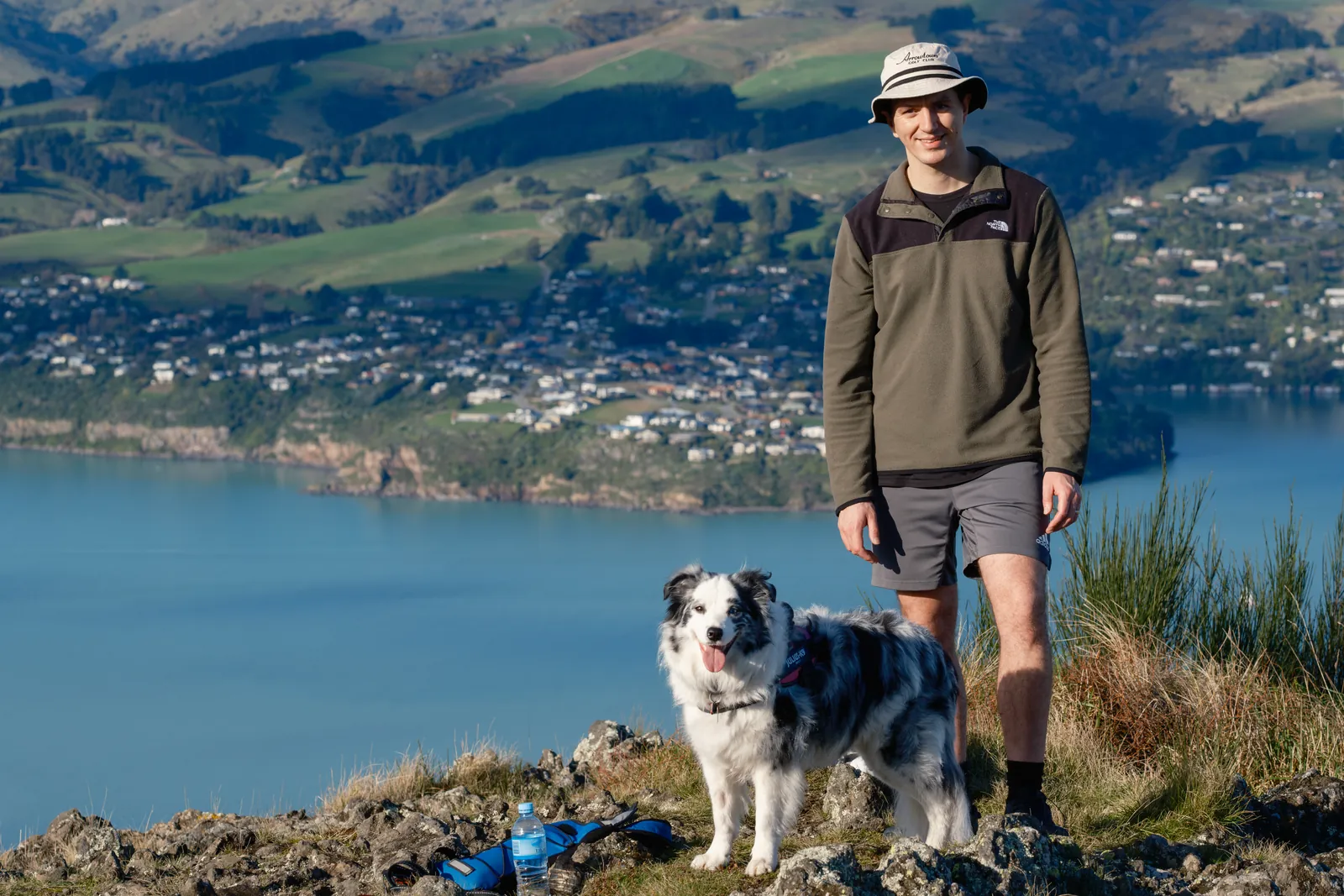 Sam Frost standing with his border collie on a hilltop above a harbour